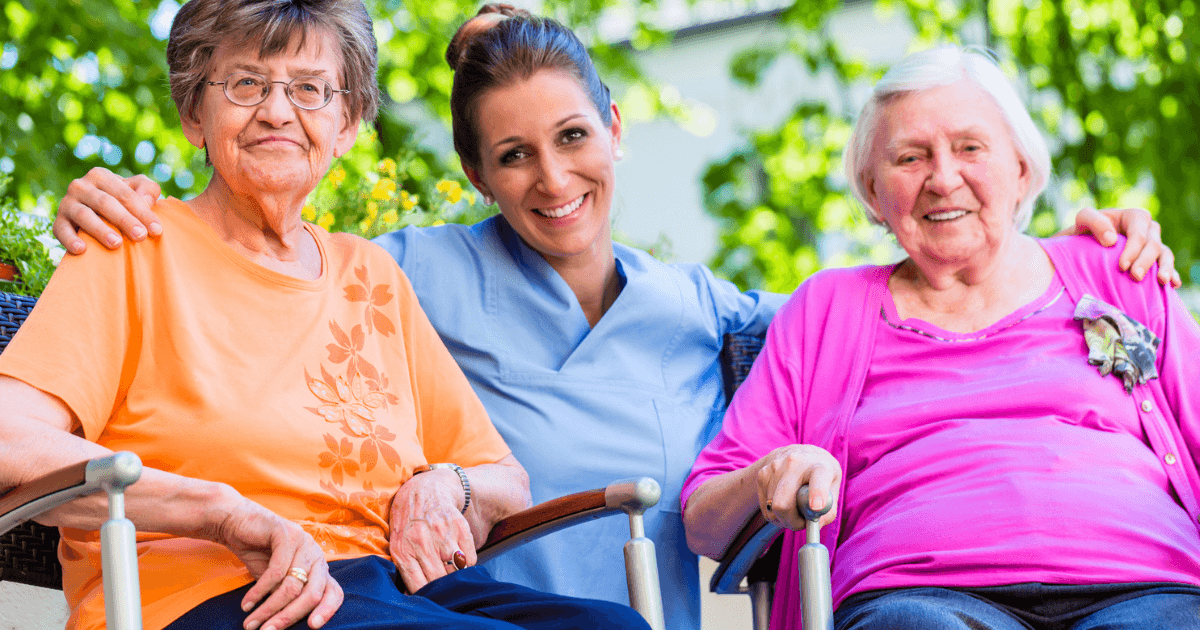 Two senior women in chairs with caregiver in between them. 